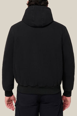 A person is shown from the back, standing against a beige background, wearing dark pants and the Hard Yakka Canvas Bomber Jacket in black.