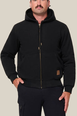 A person wearing a Hard Yakka Canvas Bomber Jacket in black with a zip-up hood, front pockets, and a small brown patch near the hem stands against a plain light beige background.