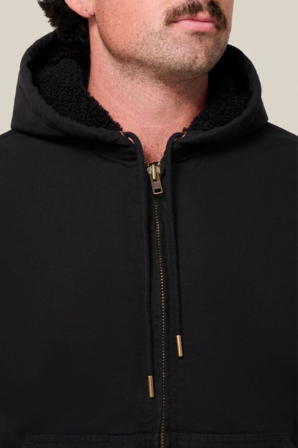 A man with a mustache wears the Hard Yakka Canvas Bomber Jacket, featuring a fleece-lined hood and gold zipper, partially zipped up against a neutral background.