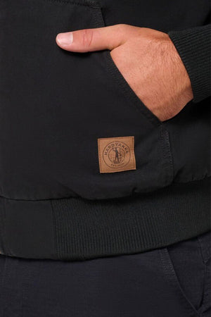 A person with a light skin tone has their right hand in the pocket of a durable Hard Yakka Canvas Bomber Jacket. A brown patch with the "Nerdyark" logo is sewn near the hem of the black jacket, which is paired with dark pants.