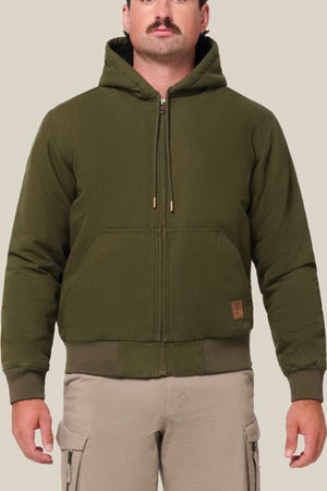 A man with a mustache wears a Hard Yakka Canvas Bomber Jacket—an olive green hooded zip-up with front pockets—and light beige cargo pants, standing against a plain light beige background.