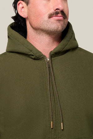 A person with a mustache wearing the Hard Yakka Canvas Bomber Jacket with sherpa lining in olive green, shown chest up against a plain beige background. The hood is up but not covering their head.