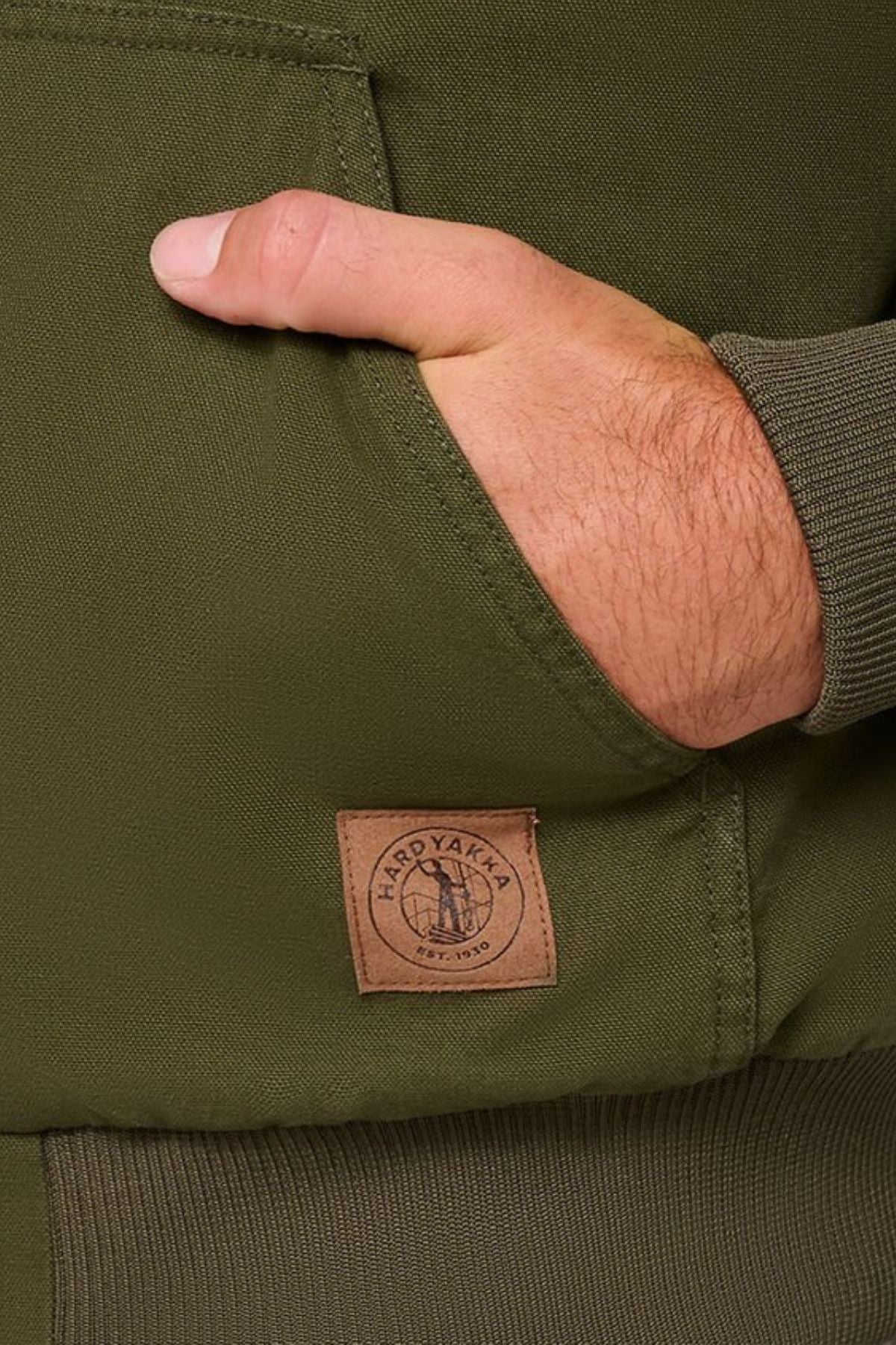 A close-up of a hand partly tucked into the pocket of an olive green Hard Yakka Canvas Bomber Jacket, featuring a logo patch near the pocket and ribbed sleeve cuff.