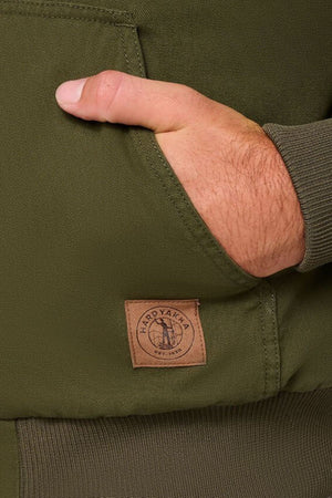 A close-up of a hand partly tucked into the pocket of an olive green Hard Yakka Canvas Bomber Jacket, featuring a logo patch near the pocket and ribbed sleeve cuff.