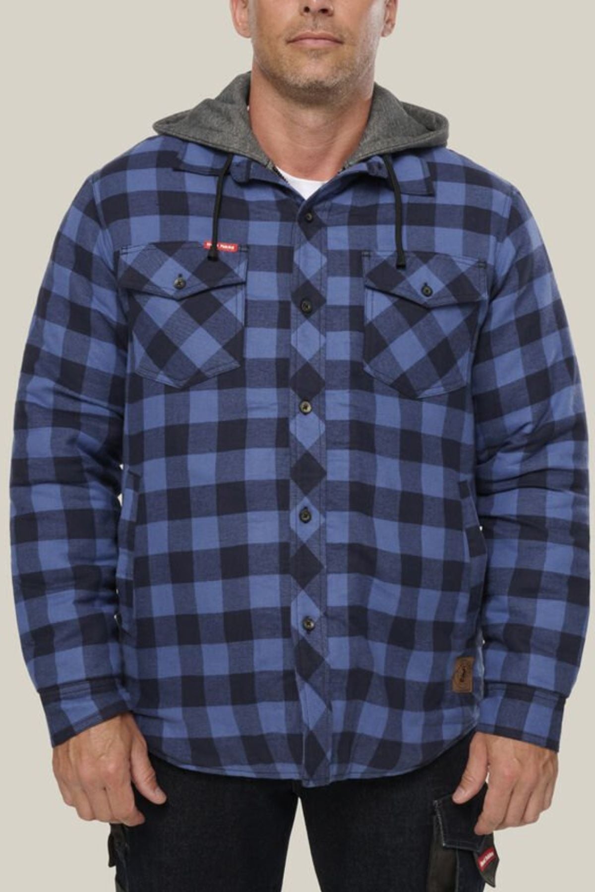 A person wearing a Hard Yakka Quilted Flannelette Shacket in blue and black plaid with a gray hood stands against a plain light background. The shacket features two chest pockets with flaps and black buttons.