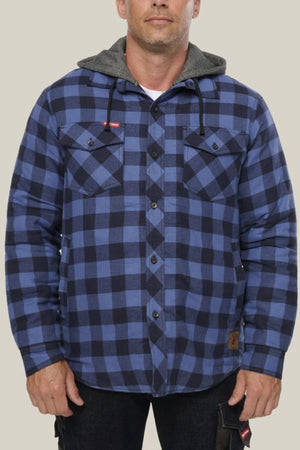 A person wearing a Hard Yakka Quilted Flannelette Shacket in blue and black plaid with a gray hood stands against a plain light background. The shacket features two chest pockets with flaps and black buttons.