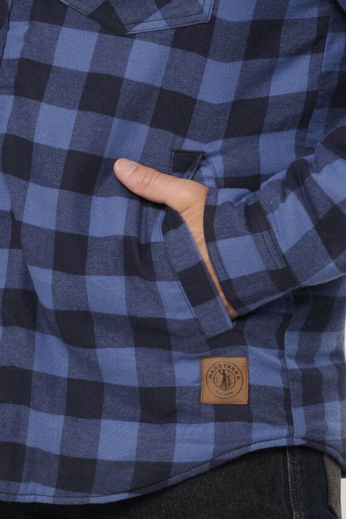 Close-up of a person wearing the Hard Yakka Quilted Flannelette Shacket in blue and black plaid, with one hand partly inside the chest pocket and a brown logo patch near the bottom hem.