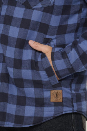Close-up of a person wearing the Hard Yakka Quilted Flannelette Shacket in blue and black plaid, with one hand partly inside the chest pocket and a brown logo patch near the bottom hem.