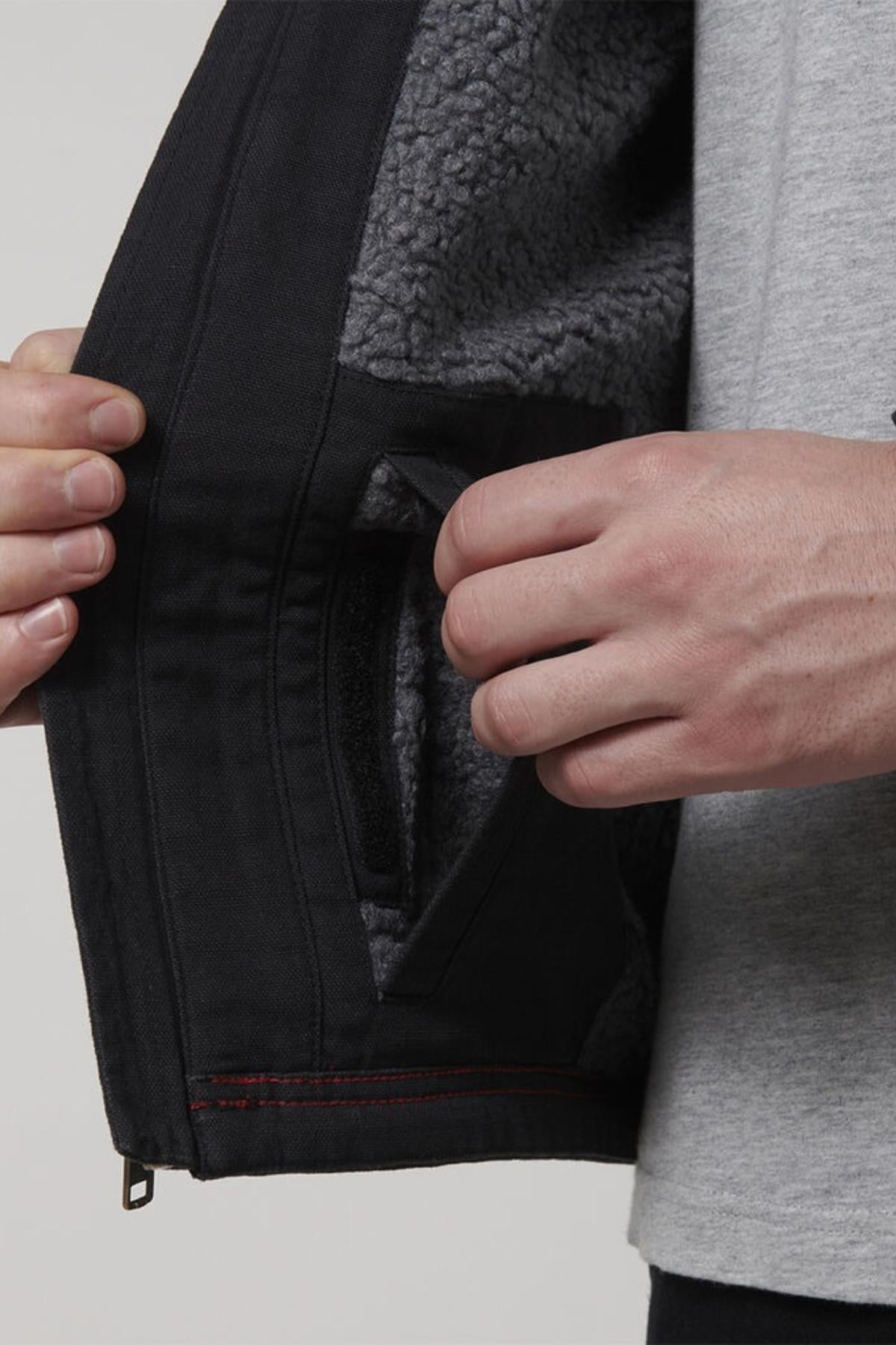 A person opens the side of a Hard Yakka Heritage Canvas Hooded Jacket, exposing a soft sherpa fleece lining. Their other hand grips near the pocket with its heavy-duty zip, while wearing a light gray shirt.