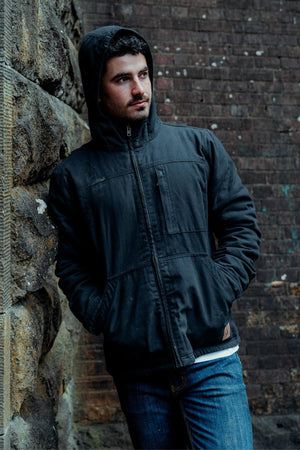 A person wears a Hard Yakka Heritage Canvas Hooded Jacket with sherpa fleece lining, standing against a stone wall. The textured brick backdrop complements the cozy warmth and style of their heavy-duty zip-up attire.