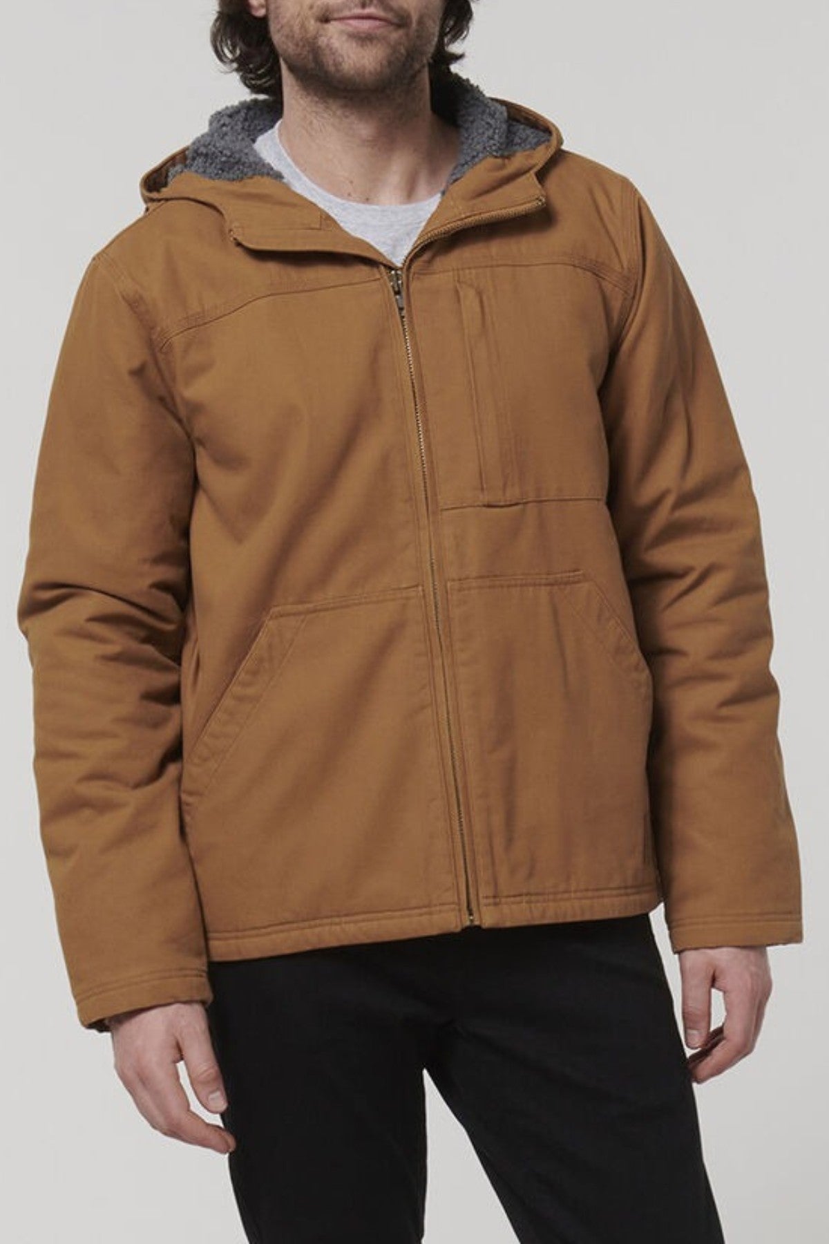 A person wearing the Hard Yakka Heritage Canvas Hooded Jacket in light brown with sherpa fleece lining, durable stitching, and front pockets stands against a plain light background, paired with black pants.