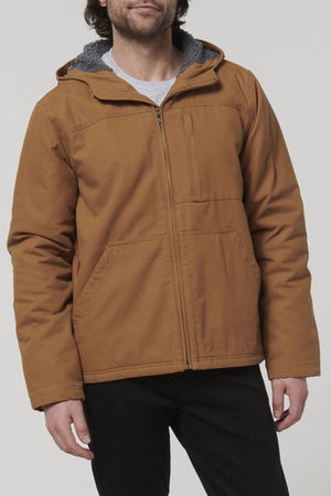 A person wearing the Hard Yakka Heritage Canvas Hooded Jacket in light brown with sherpa fleece lining, durable stitching, and front pockets stands against a plain light background, paired with black pants.