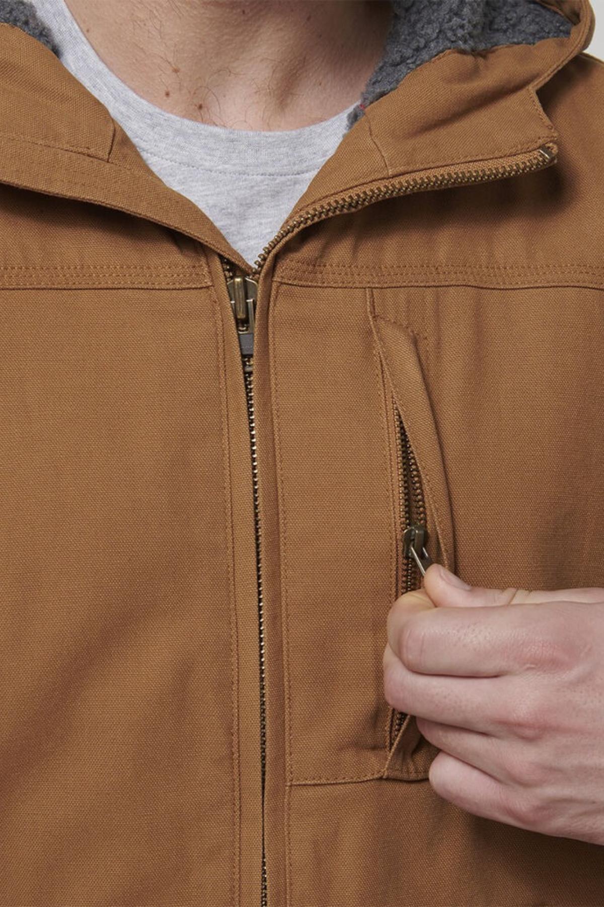 A person wearing the Hard Yakka Heritage Canvas Hooded Jacket with a sherpa fleece-lined collar is unzipping a small chest pocket. The jacket, known for its durable stitching, is layered over a light gray shirt.
