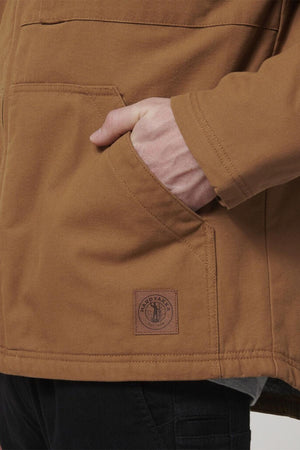 A person wears the Hard Yakka Heritage Canvas Hooded Jacket, featuring durable stitching and a visible patch near the pocket, with their right hand in the pocket, styled with black pants.