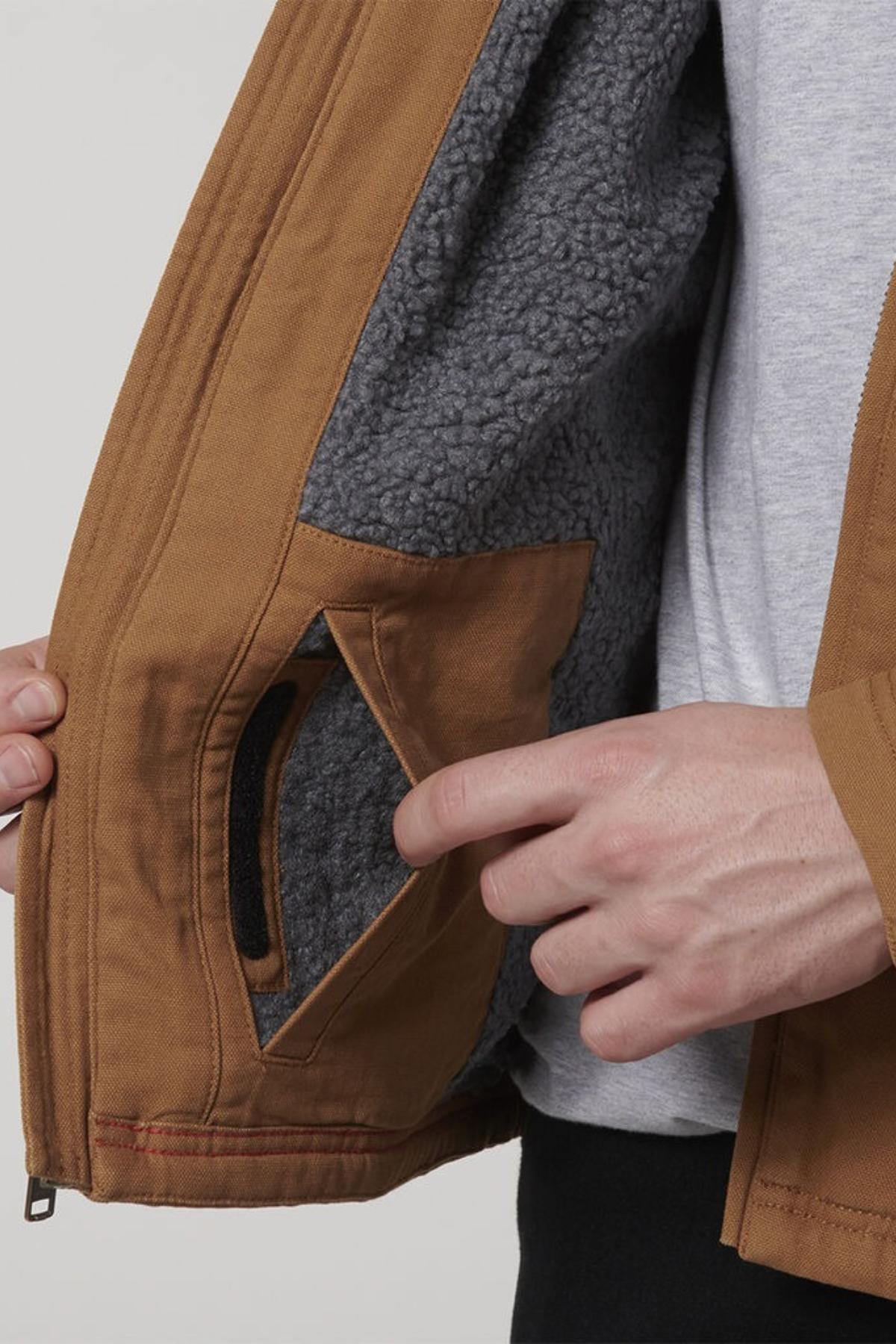 A person opens a Hard Yakka Heritage Canvas Hooded Jacket, revealing its cozy Sherpa fleece lining and an inner pocket; only their torso and hands are visible, showing durable stitching throughout and a light gray shirt underneath.
