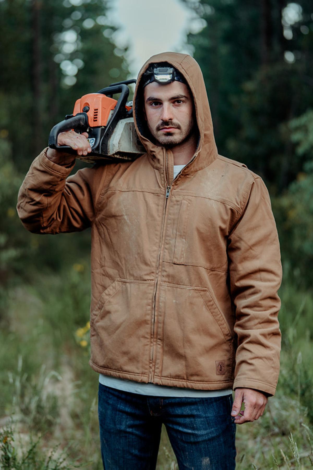 A man in the Hard Yakka Heritage Canvas Hooded Jacket stands outdoors in a wooded area, holding a chainsaw on his shoulder and wearing a headlamp, surrounded by trees and greenery.