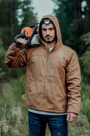 A man in the Hard Yakka Heritage Canvas Hooded Jacket stands outdoors in a wooded area, holding a chainsaw on his shoulder and wearing a headlamp, surrounded by trees and greenery.