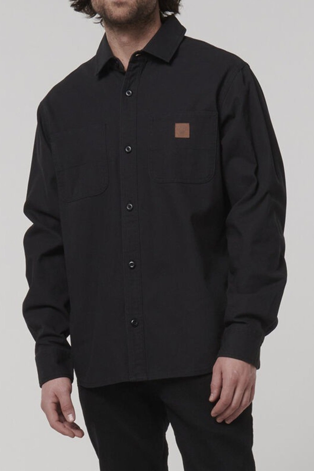 A man wears the Hard Yakka Heritage Canvas Shirt in black, featuring long sleeves and two chest pockets, standing against a plain light background with his face partially out of frame.