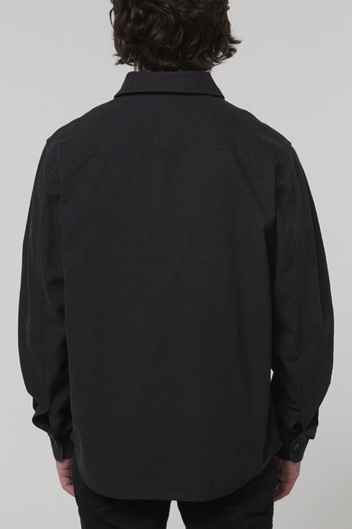 A person with medium-length dark hair is shown from the back, wearing a Hard Yakka Heritage Canvas Shirt with long sleeves, blending style and functionality against a plain light background.