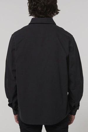 A person with medium-length dark hair is shown from the back, wearing a Hard Yakka Heritage Canvas Shirt with long sleeves, blending style and functionality against a plain light background.