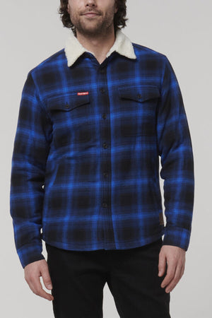 A man stands against a plain background wearing a Hard Yakka Heritage Trucker Sherpa Jacket in blue and black plaid. This regular fit jacket boasts a cozy sherpa collar, button-up closure, and practical chest pockets, merging functionality with style.