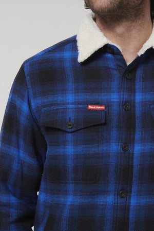 A person is wearing a Hard Yakka Heritage Trucker Sherpa Jacket in blue and black plaid, featuring button closures, a sherpa collar, and a regular fit. The jacket has a chest pocket with a red label; the individual's face remains unseen.