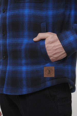 Wearing a blue and black plaid shirt with black buttons and a brown leather patch, paired with black pants, the outfit mirrors the understated style of the Hard Yakka Heritage Trucker Sherpa Jacket.