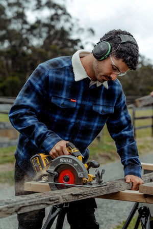 Wearing protective eyewear and earmuffs, a person cuts a wooden plank with a circular saw while donning a Hard Yakka Heritage Trucker Sherpa Jacket. In the background, tall trees are silhouetted against a cloudy sky.