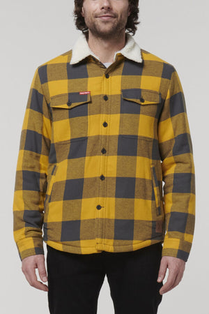 A man wears the Hard Yakka Heritage Trucker Sherpa Jacket—a yellow and black checkered 100% cotton flannel with a white sherpa collar, button front, and pockets—standing against a plain light gray background.