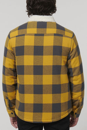 A person with dark hair is seen from behind, wearing the Hard Yakka Heritage Trucker Sherpa Jacket in yellow and black check with a white sherpa collar and black pants, against a plain light background.