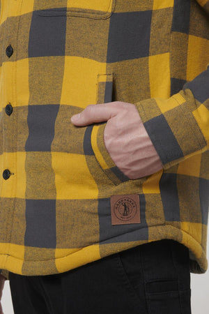 A person wears the Hard Yakka Heritage Trucker Sherpa Jacket—a yellow and gray plaid, 100% cotton button-up with a Sherpa collar, logo patch near the hem, and black pants. One hand is in their pocket.