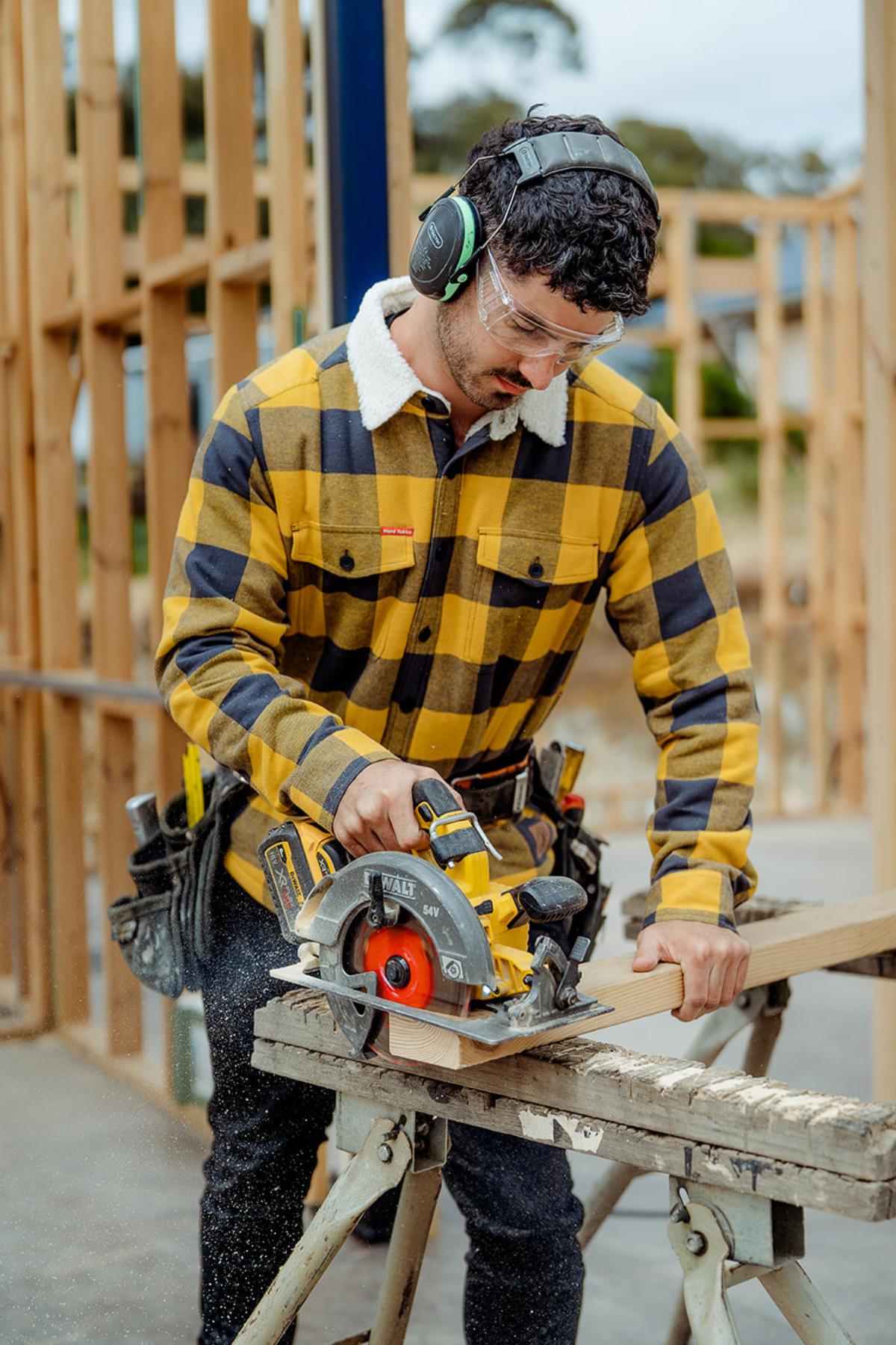 A man in a Hard Yakka Heritage Trucker Sherpa Jacket, safety glasses, and earmuffs uses a circular saw to cut wood on a construction site with wooden framing and sawdust visible in the background.