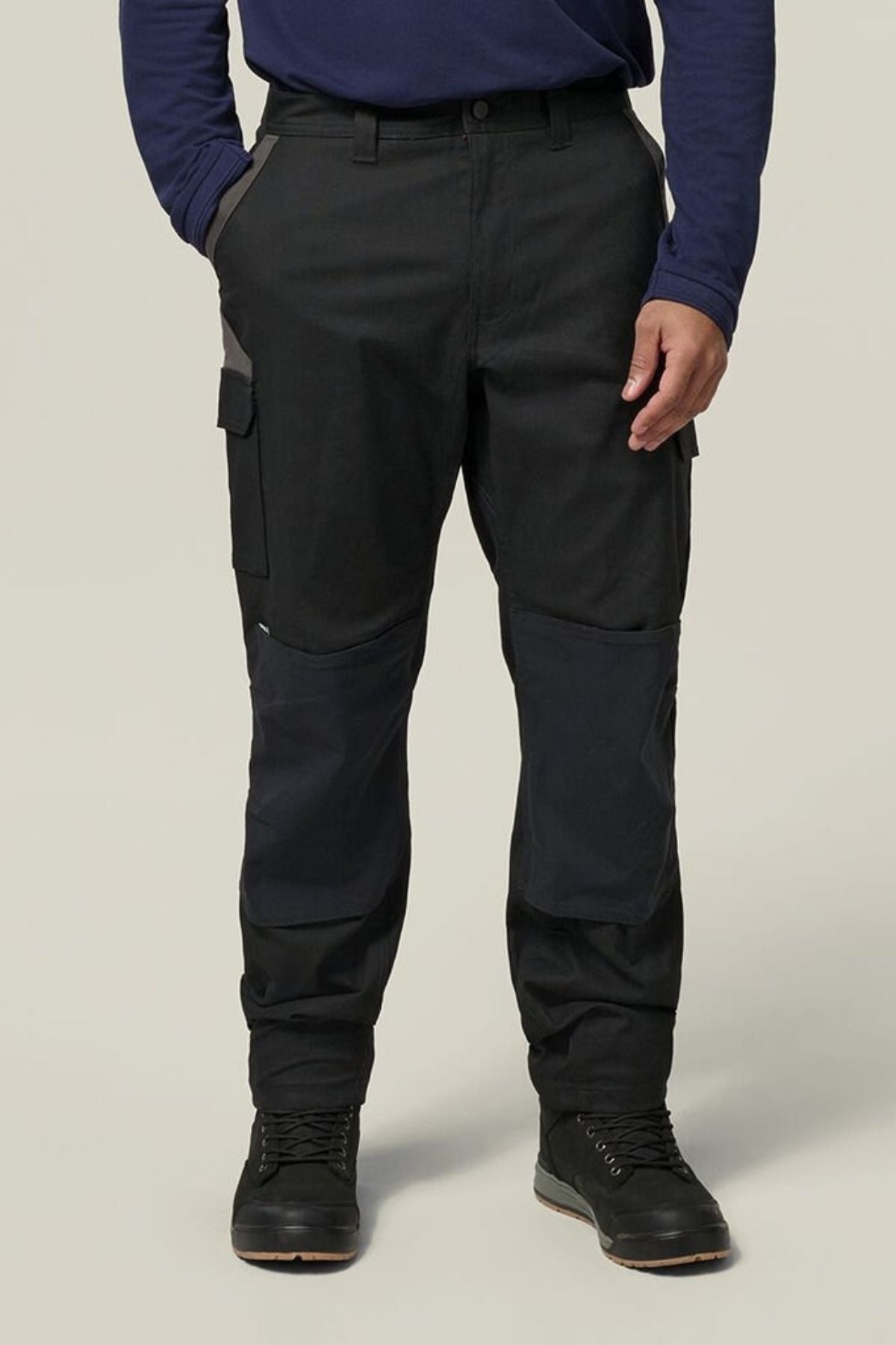 A person wearing Hard Yakka Legends Slim Stretch Work Pant in black, a long-sleeve navy blue shirt, and black boots stands with hands in pockets against a plain light background.