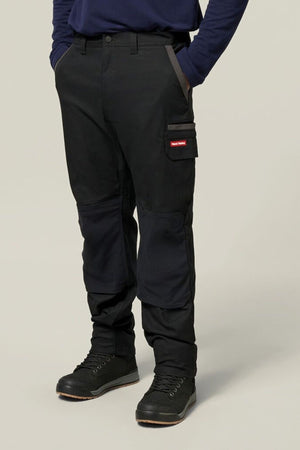 A person in Hard Yakka Legends Slim Stretch Work Pant with knee patches, red and white side pocket patch, black boots, and a navy long-sleeve shirt. Hands in pockets, standing against a plain light gray background.