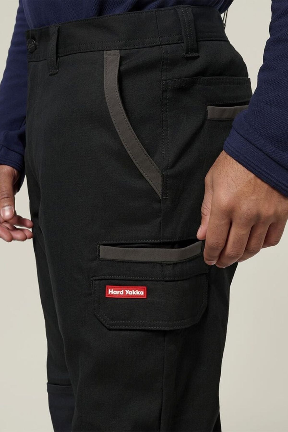 A person wearing Hard Yakka Legends Slim Stretch Work Pant with gray accents, multiple pockets including one with a red "Hard Yakka" label, and a navy long-sleeve shirt; only the lower torso and hand are visible.
