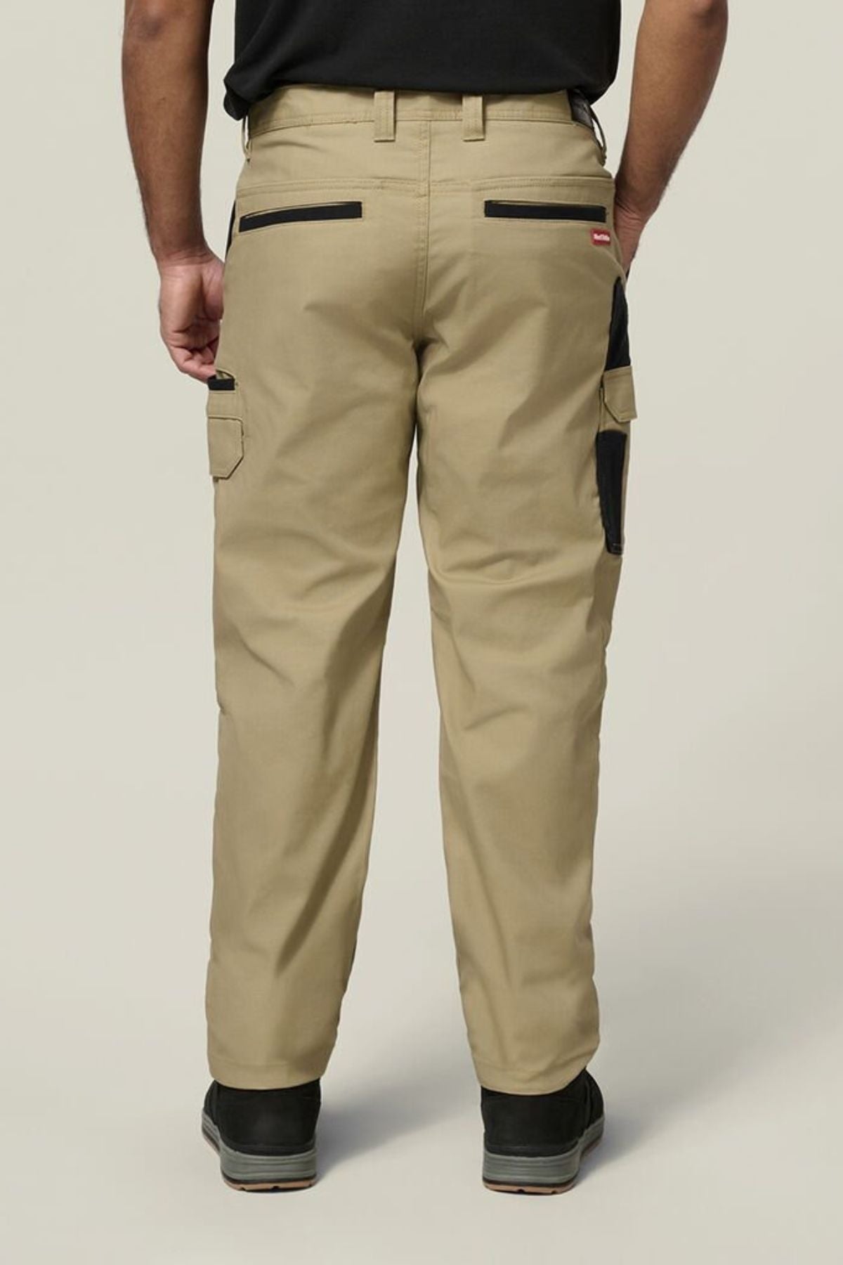 A person stands facing away from the camera, highlighting the back of the Hard Yakka Legends Slim Stretch Work Pant with utility pockets and black accents, paired with a black shirt and black shoes.