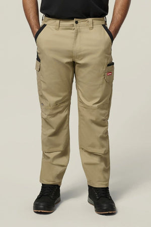 A person wearing tan Hard Yakka Legends Slim Stretch Work Pant with multiple utility and cargo pockets, paired with black boots, stands facing forward with hands in the front pockets against a plain light-colored background.