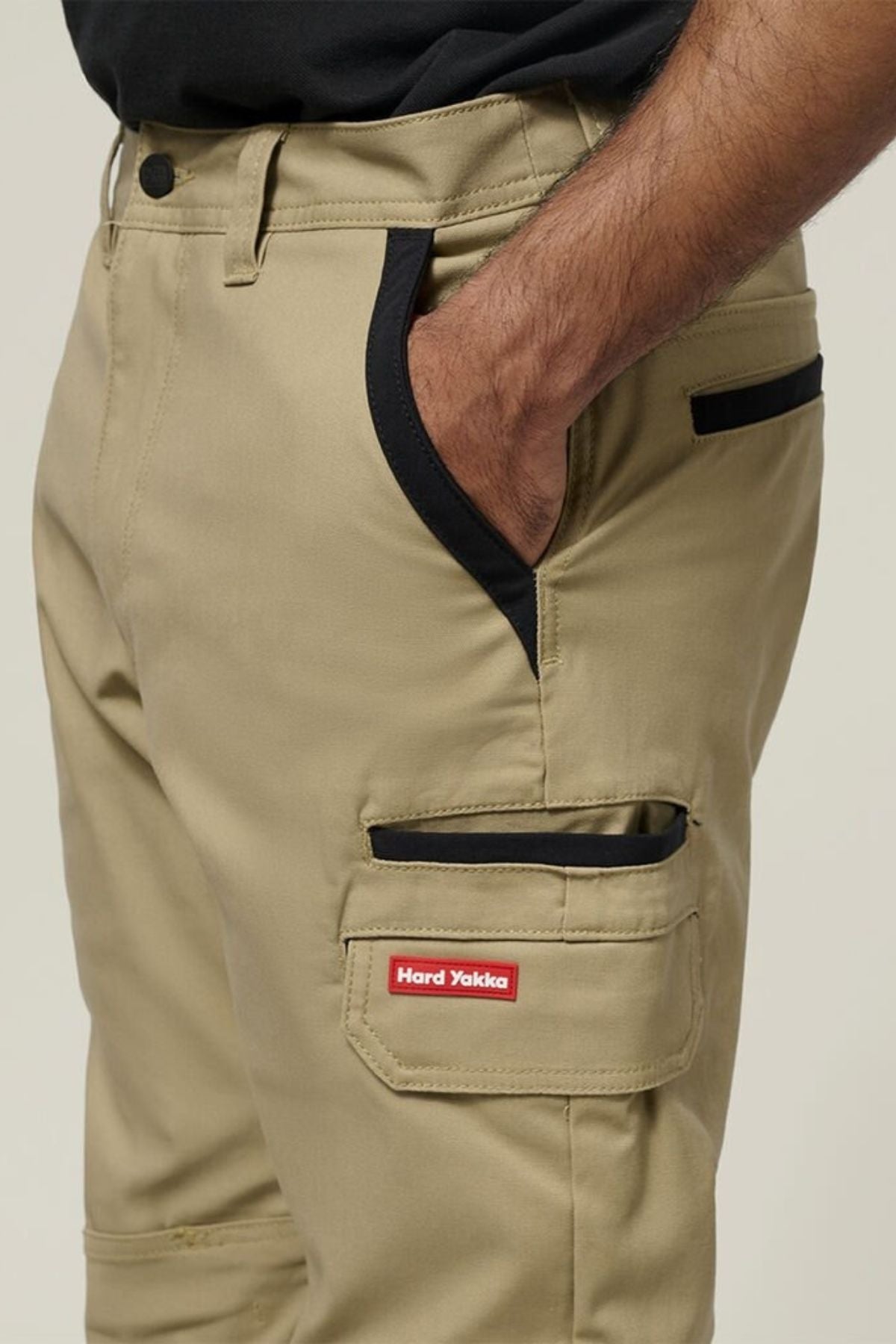 A person wears Hard Yakka Legends Slim Stretch Work Pant in tan with black trim, a red label, utility pockets, and a black-collared pocket detail. Their right hand is in the front pocket, paired with a black shirt.