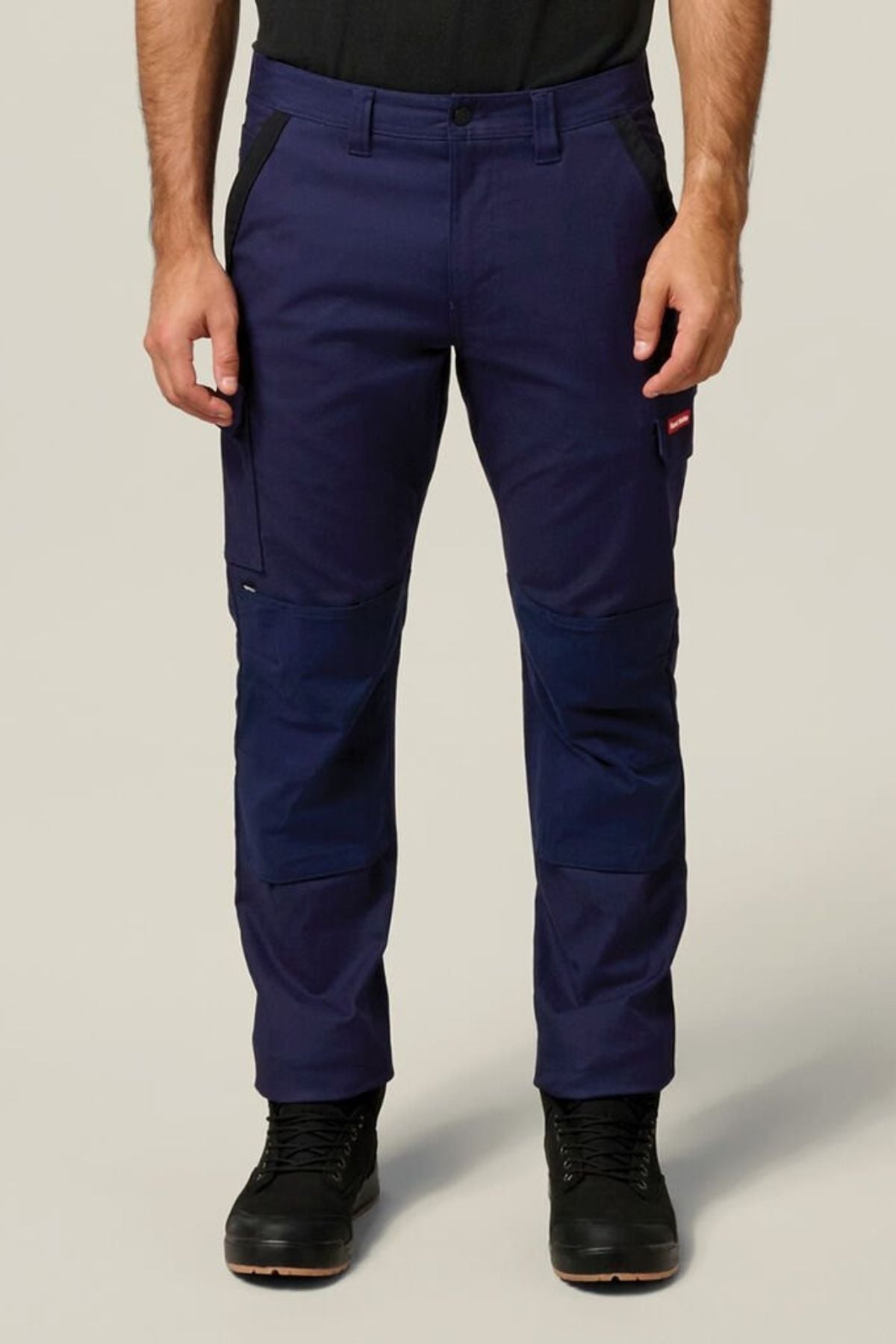 A person wearing Hard Yakka Legends Slim Stretch Work Pant in navy with utility and spacious side pockets, reinforced knees, plus black boots and a black shirt stands against a plain light background.