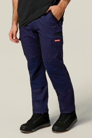 A person wearing Hard Yakka Legends Slim Stretch Work Pant in dark blue, featuring reinforced knees and utility pockets, along with black shoes and a black shirt with one hand in the pocket, stands against a plain light background.