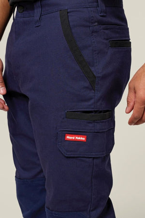 A close-up shows a person wearing the Hard Yakka Legends Slim Stretch Work Pant in dark blue with black trim, featuring multiple utility pockets and a flap pocket labeled "Hard Yakka" in red and white. The person's hand rests by their side.