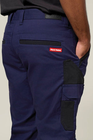 A person wears Hard Yakka Legends Slim Stretch Work Pant in navy blue with black reinforced utility pockets and a red "Hard Yakka" label on the back pocket. Their right arm is visible against a neutral background.