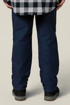 A person stands facing away, wearing a black-and-white checkered shirt, Hard Yakka Moleskin Plain Front Heavy Duty Work Pant in dark blue, and black shoes. The background is plain and light-colored.