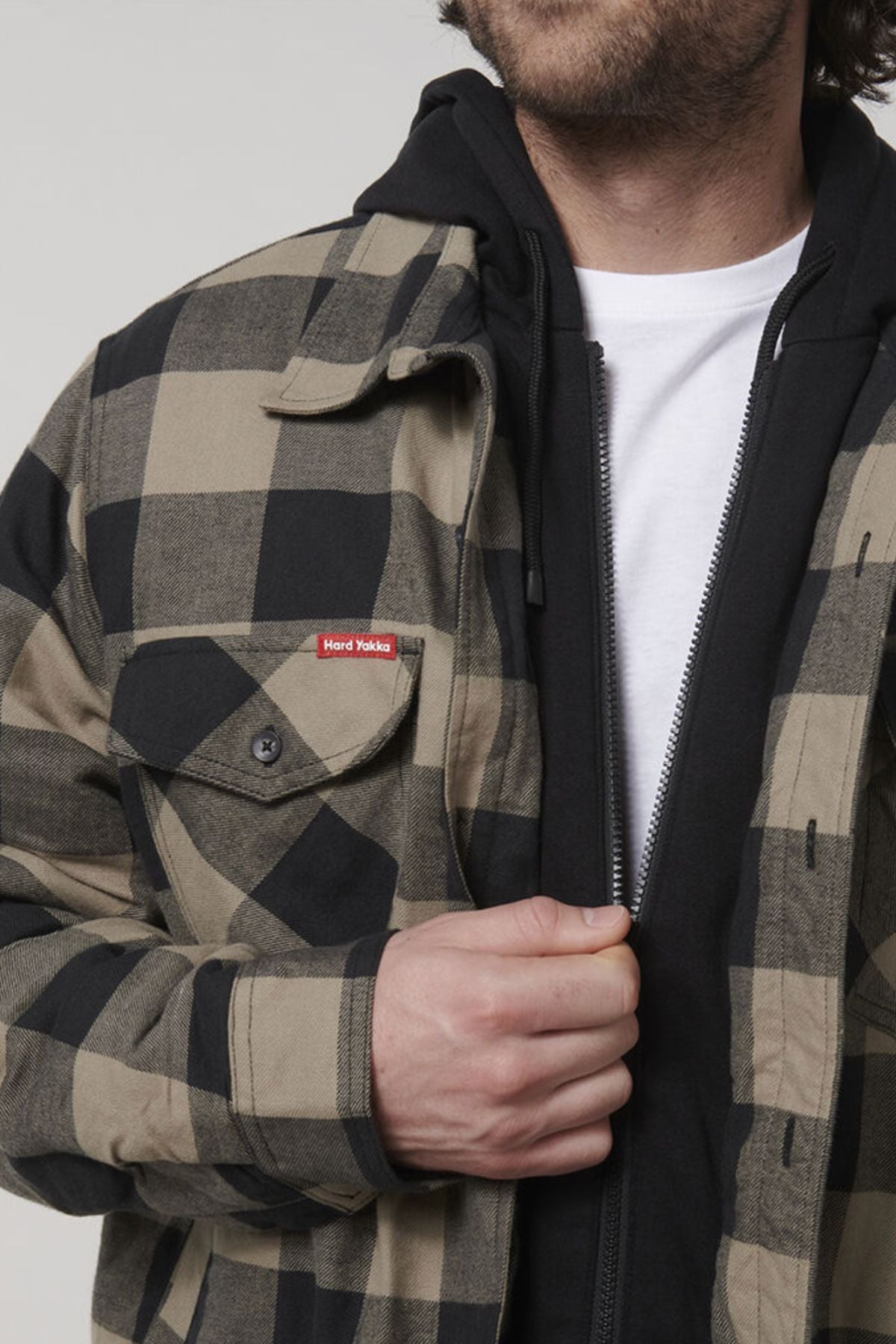 A person is wearing a Hard Yakka Quilted Flannelette Shacket in a black and beige checkered pattern over a black hoodie and white shirt. Their hand is partially adjusting the shacket, revealing a small red tag reading "Hard Yakka" on the left chest pocket. This comfortable, hard-wearing shacket stands out against the plain, neutral background.