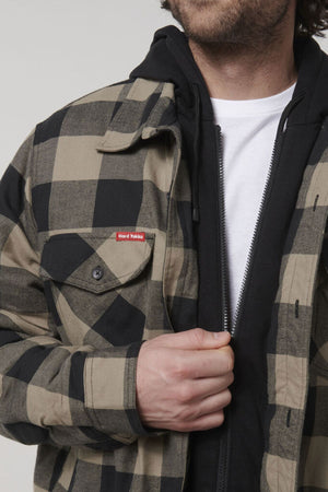 A person is wearing a Hard Yakka Quilted Flannelette Shacket in a black and beige checkered pattern over a black hoodie and white shirt. Their hand is partially adjusting the shacket, revealing a small red tag reading "Hard Yakka" on the left chest pocket. This comfortable, hard-wearing shacket stands out against the plain, neutral background.