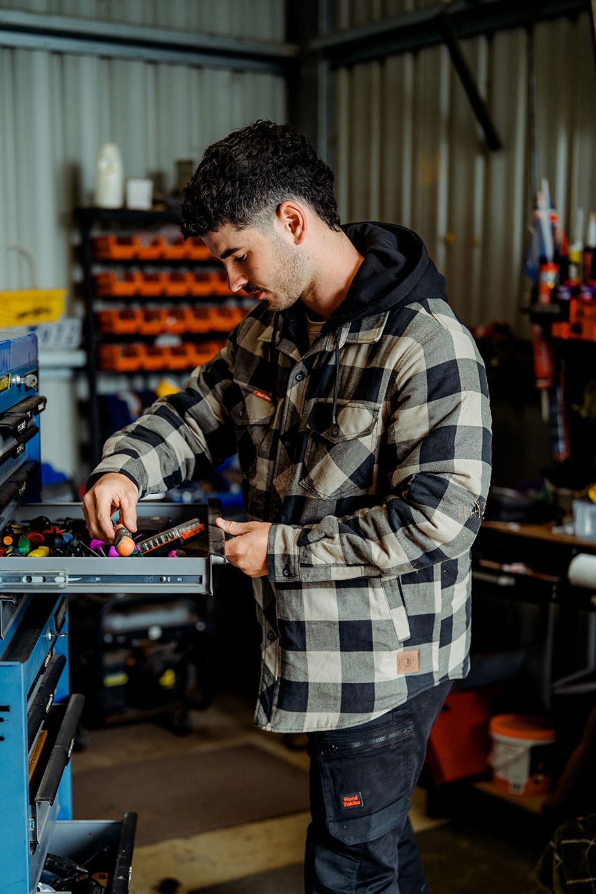 A person wearing a Hard Yakka Quilted Flannelette Shacket and dark pants organizes tools in a drawer inside a well-equipped workshop, with various Yakka tools, shelves, and storage bins visible in the background.
