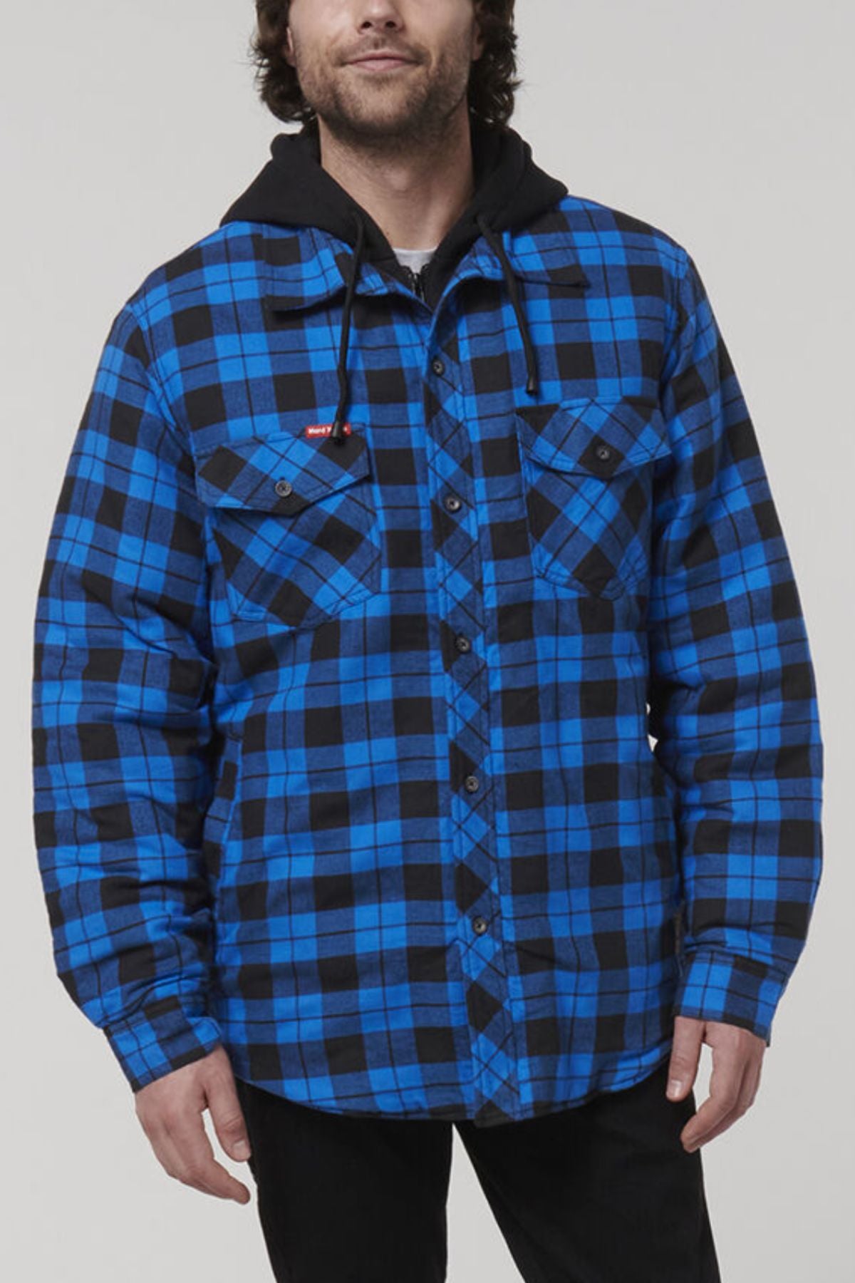 A person wearing the Hard Yakka Quilted Flannelette Shacket, featuring a blue and black plaid pattern, two chest pockets, and a black hood, stands against a plain background. Only the upper part of their body is visible.