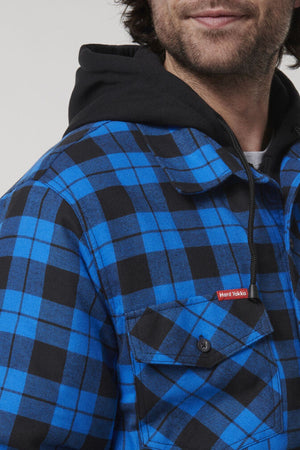 A person wearing a vibrant blue and black plaid Hard Yakka Quilted Flannelette Shacket over a black hoodie is shown from the shoulders up, smiling slightly. The shacket features a red label on the chest pocket. The background is plain and light-colored.