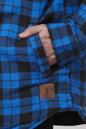 Close-up of a person wearing the Hard Yakka Quilted Flannelette Shacket in a blue and black checkered pattern, with their hand in the pocket. A brown patch featuring the text "Hard Yakka" and a logo is stitched near the hem of this durable jacket.
