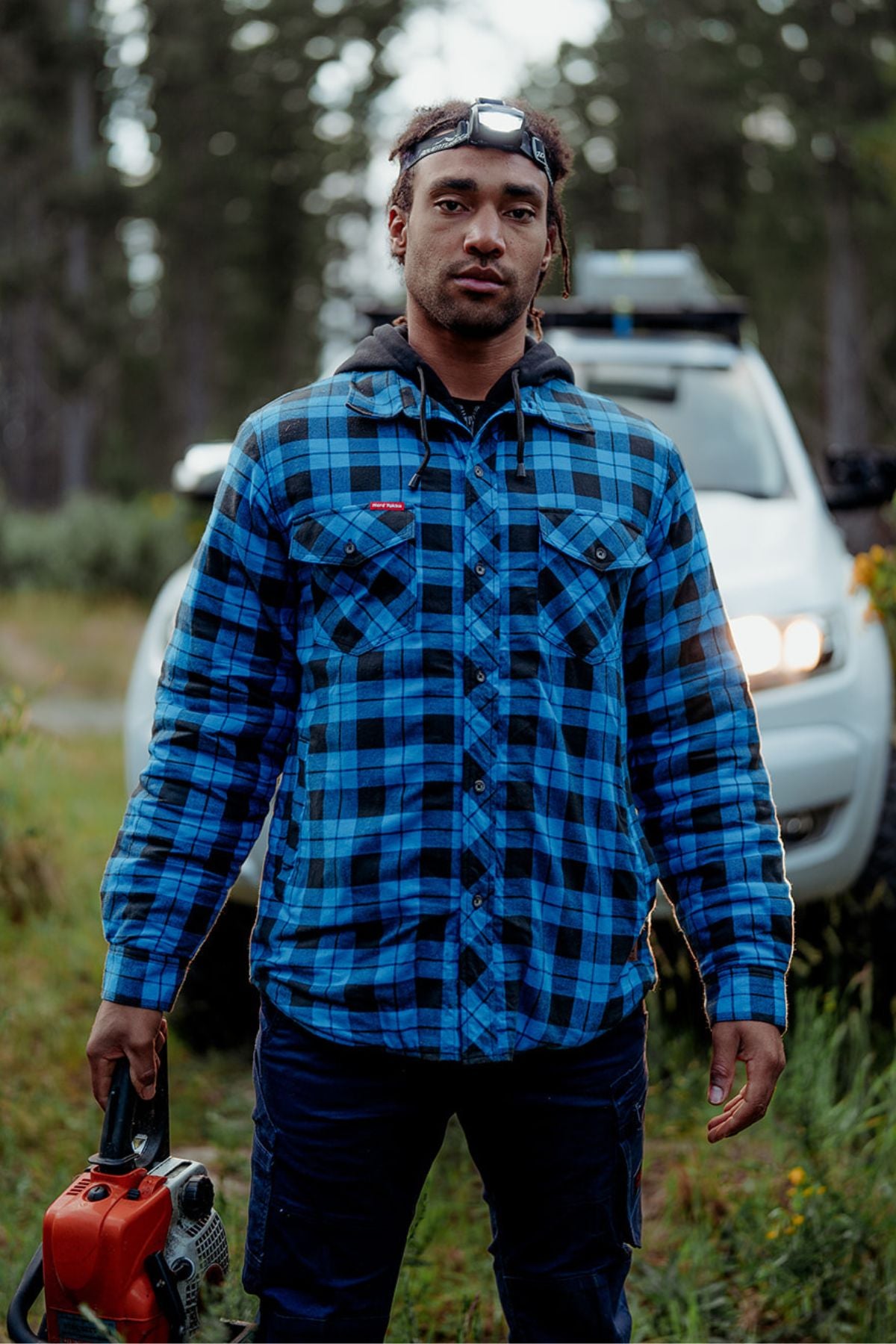 A person in a blue and black plaid shirt, Hard Yakka Quilted Flannelette Shacket, and headlamp stands in front of a white SUV, holding a chainsaw in a wooded area. The forest background is slightly blurred, and the vehicle's headlights are on.